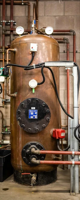 A large copper hot water storage cylinder in a building services training facility, with multiple pipe connections, pressure gauges, and insulation components. A large copper hot water storage cylinder in a building services training facility, with multiple pipe connections, pressure gauges, and insulation components.